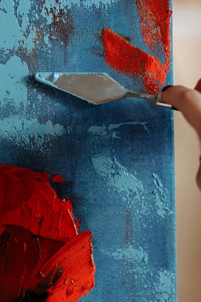 Close-up of palette knife applying vibrant red acrylic paint on a textured blue canvas.